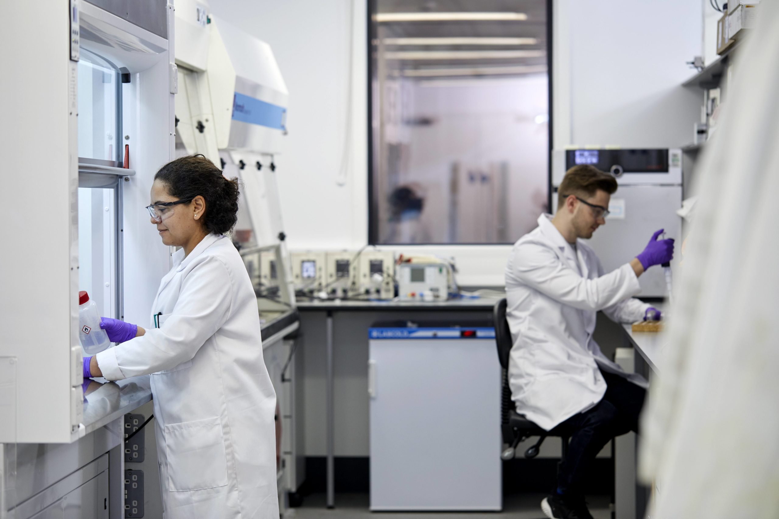 A female scientist stands working in a fume cupboard whilst her male colleague sits a workbench in laboratory