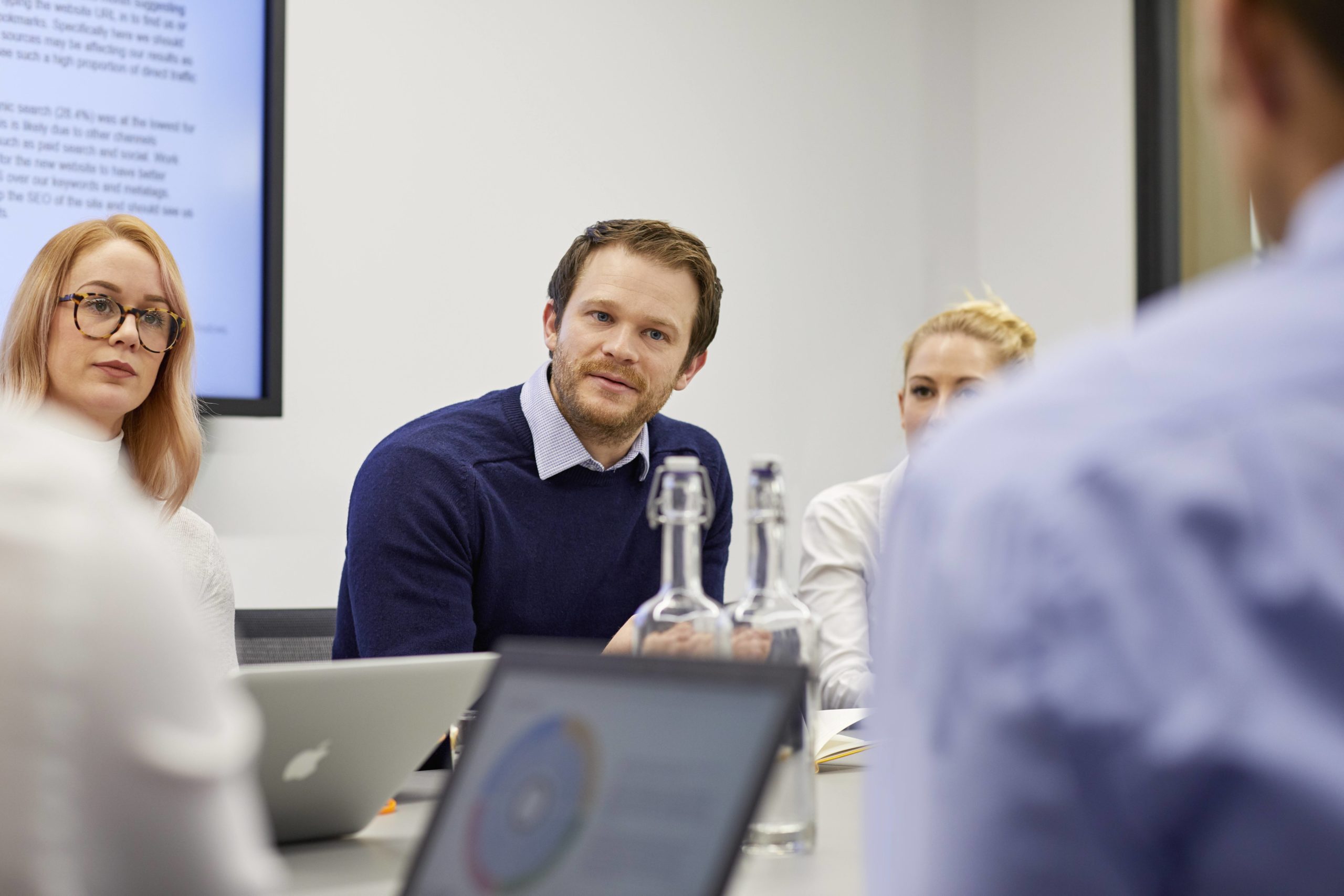 Male in centre of image talking during business meeting, female colleagues sit to left and right of him with laptops in front of them