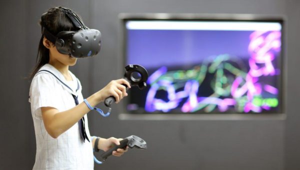 Image of student in left of frame standing with black VR headset on and holding motion devices in either hands
