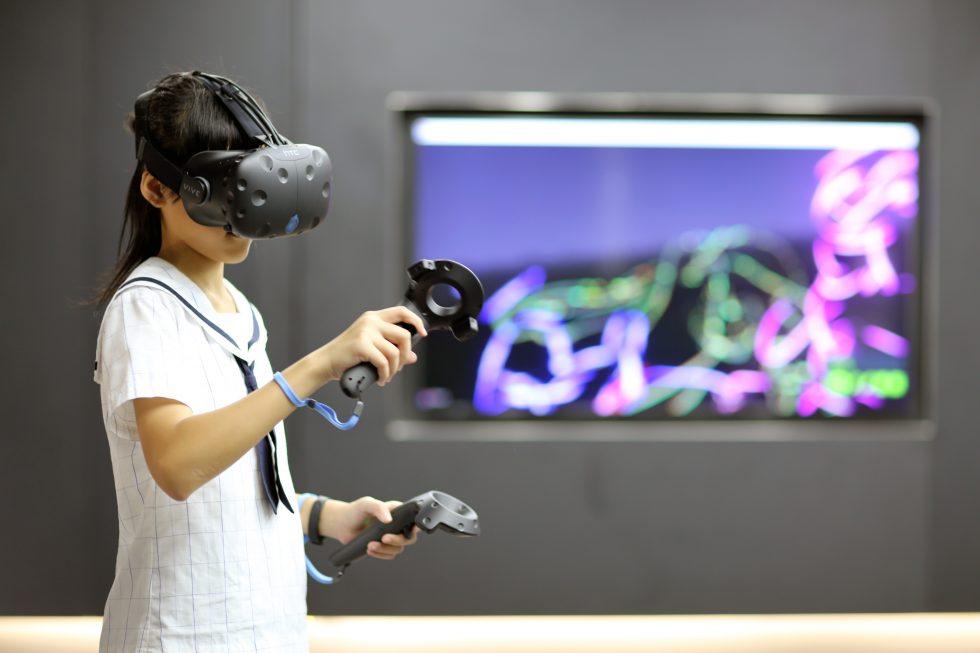 Image of student in left of frame standing with black VR headset on and holding motion devices in either hands