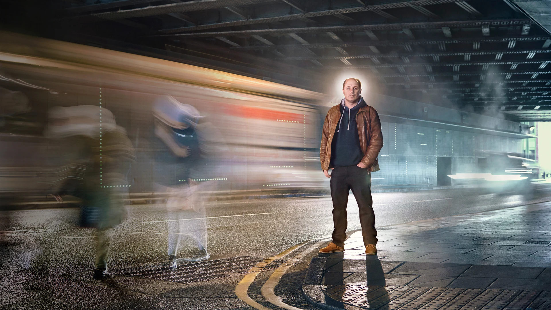 David McKee stood in tunnel under road bridge in Leeds. traffic and people around him are blurred.