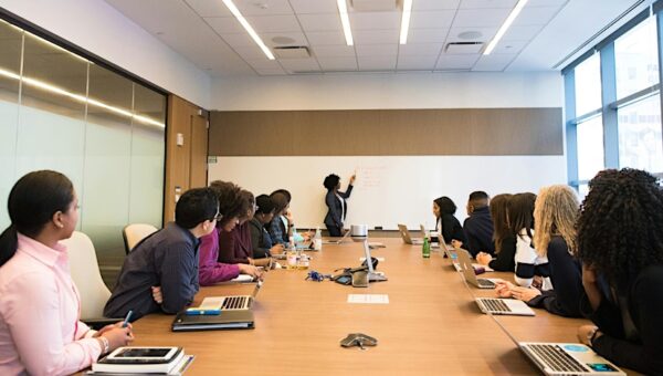 People sat at a table watching a woman write on a whiteboard.