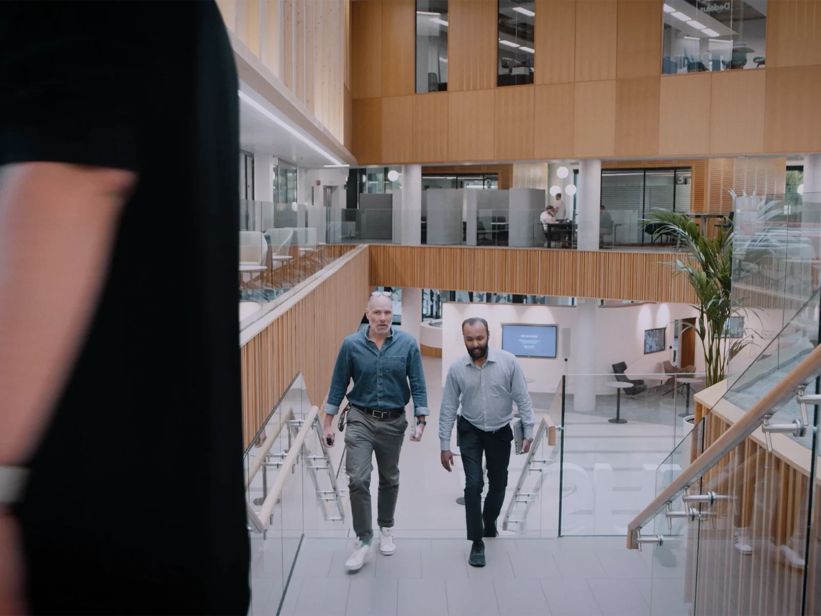 Two men walk up the Nexus atrium stairs, one wears a blue denim shirt and glasses, the other wears a pale blue shirt. Both carry laptops. In the foreground, out of focus, a person in a black tshirt walks past.