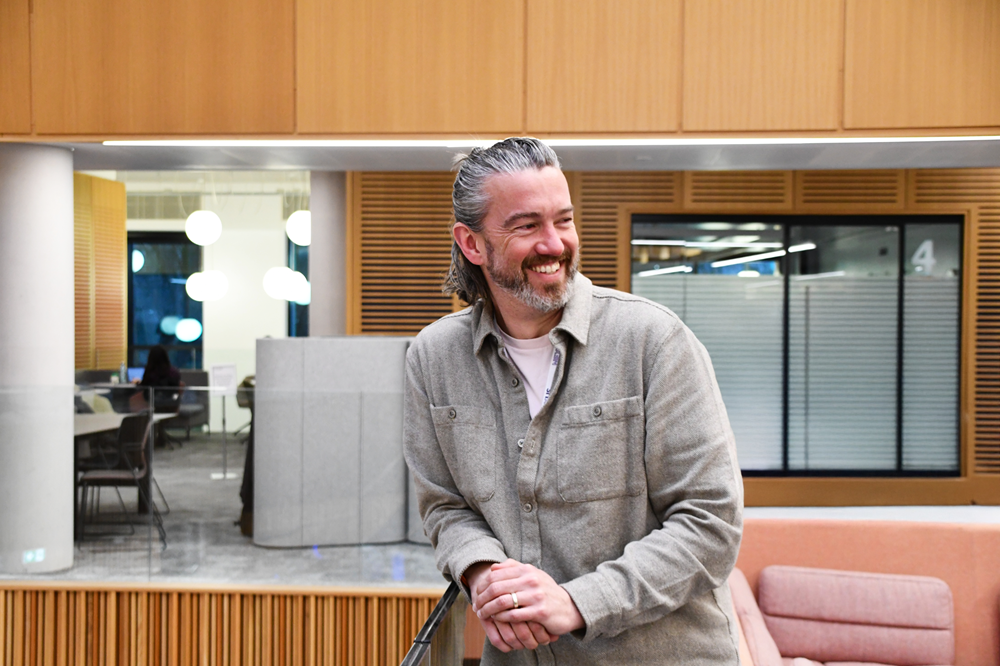 Man in a shirt stood leaning on a glass half-wall, looking out and smiling