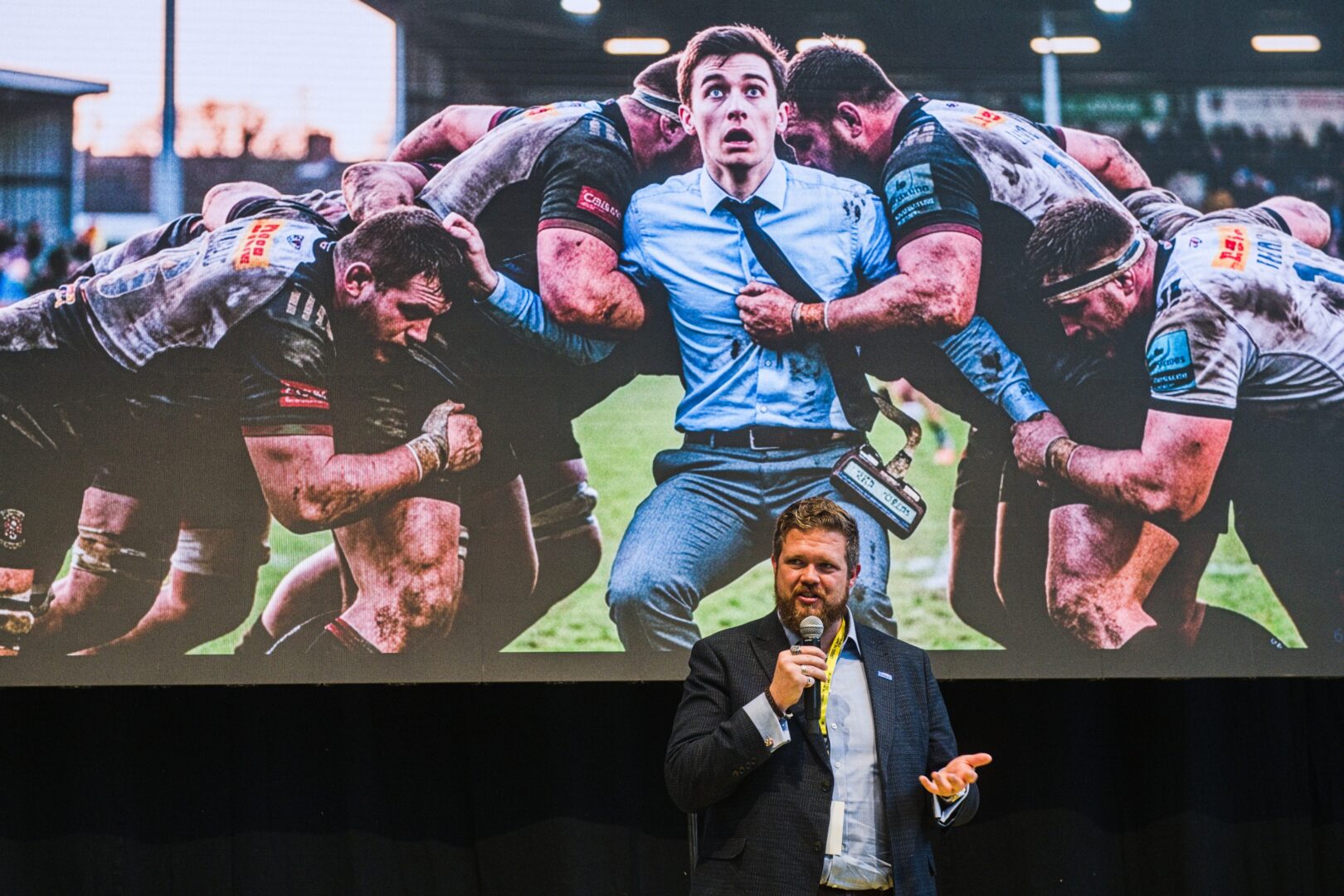 Dennis Dokter holds a microphone. Behind him is a presentation showing a man being tackled by rugby players.