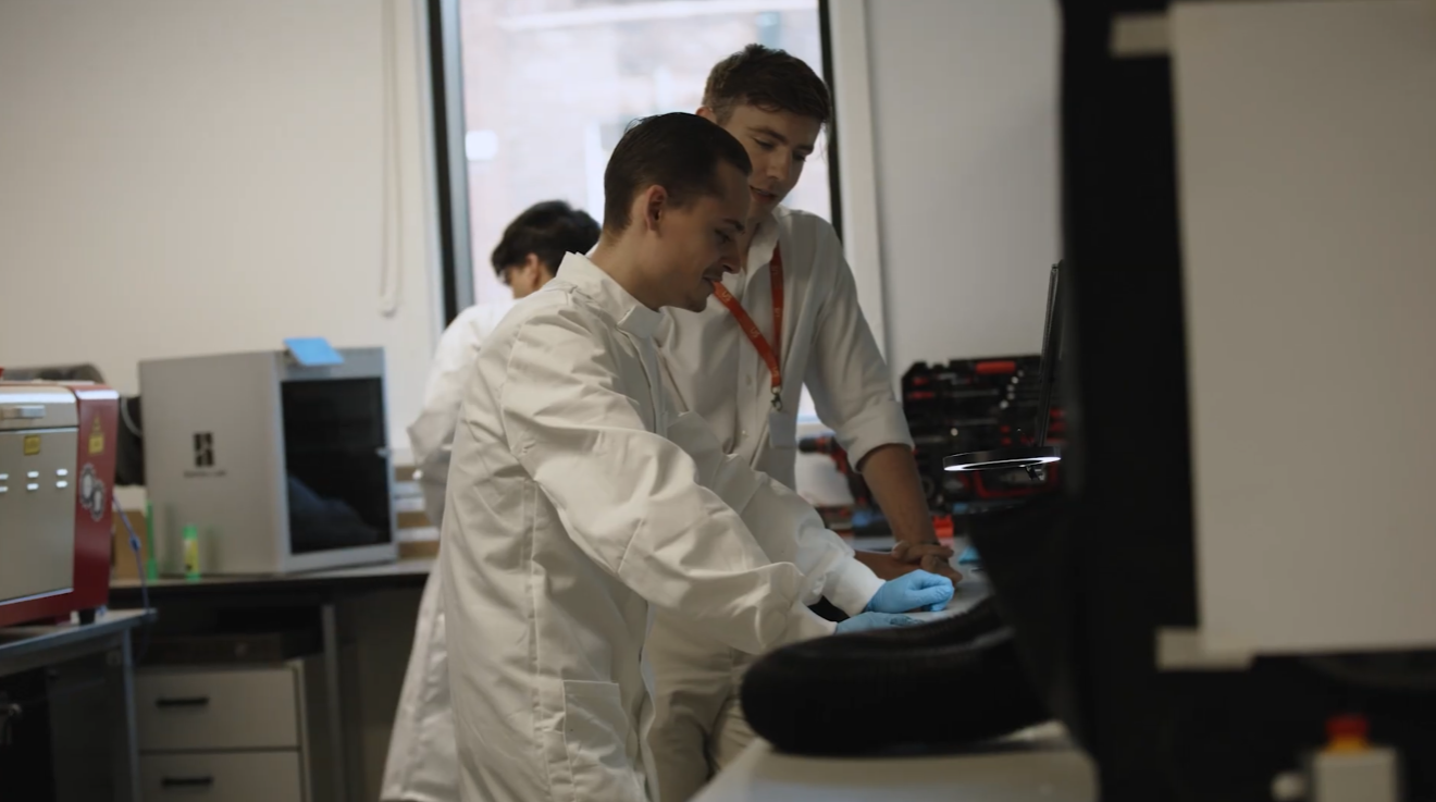Two men stood in a lab. One has a white lab coat on.